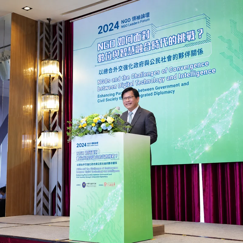2024 NGO領袖論壇講者於花卉講台後演講，背景螢幕呈現數位與智慧融合時代的挑戰及夥伴關係主題