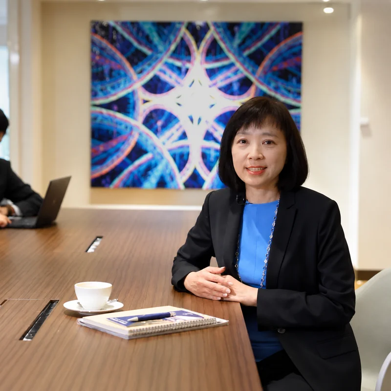 身穿黑色西裝與藍色上衣的受訪者坐在木質會議桌旁，桌上擺放茶杯與筆記本，背景為彩色藝術畫作