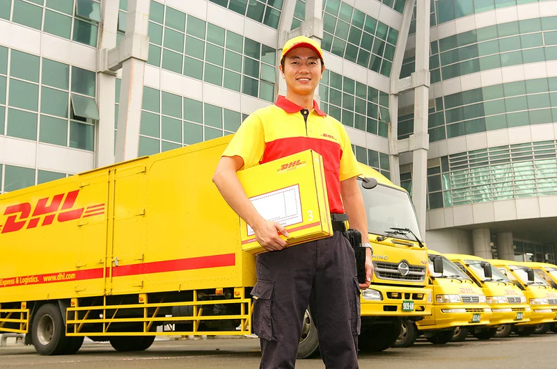 DHL配送人員抱著包裹站在黃色貨車與廂型車隊前，背景為物流中心