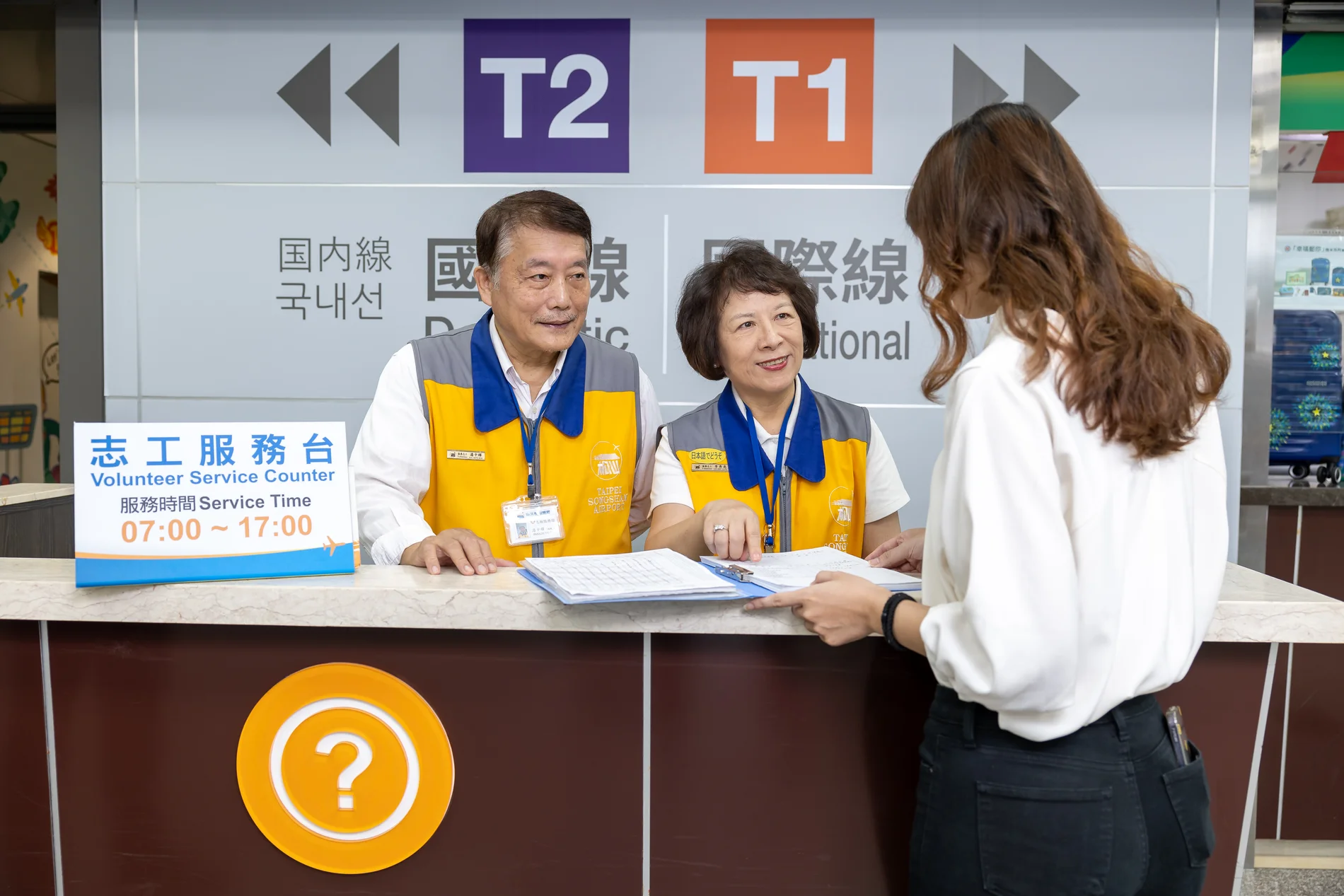 松山機場兩位志工在服務台與旅客查看資料,後方可見航廈指示標誌